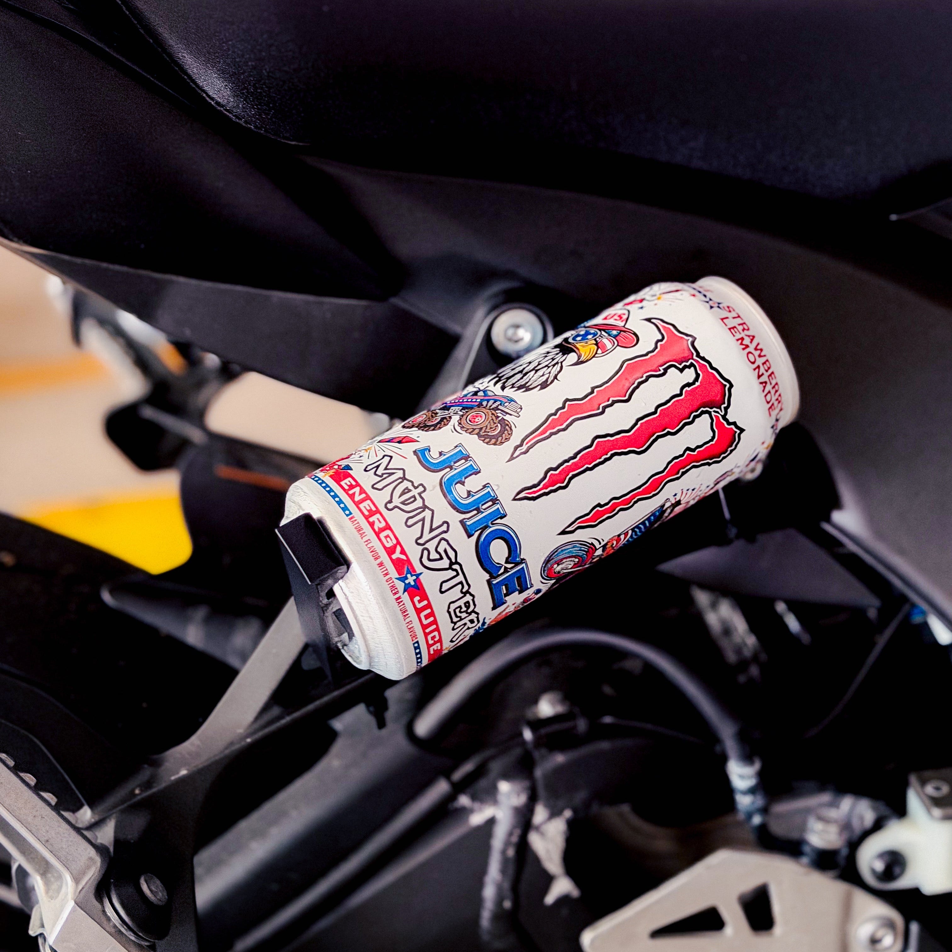 A close-up shot of a Monster Energy drink can mounted on the side of a black motorcycle, with the Monster claw logo and 'Juice' branding clearly visible.