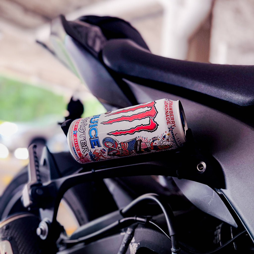 A Monster Energy drink can resting on the black handlebar of a motorcycle in an outdoor setting.